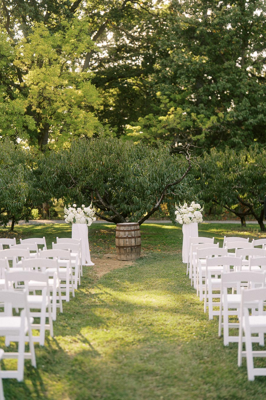 Outdoor wedding ceremony setup in a lush garden with white chairs arranged in rows facing a wooden barrel altar, surrounded by trees and decorated with white floral arrangements.