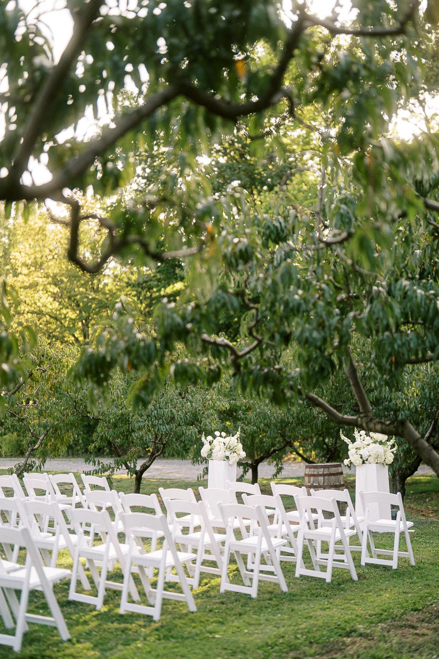 Outdoor wedding setup with rows of white chairs arranged on a grassy lawn, surrounded by lush green trees. Two white stands adorned with white flowers enhance the serene and romantic atmosphere. Ideal for garden wedding venues.