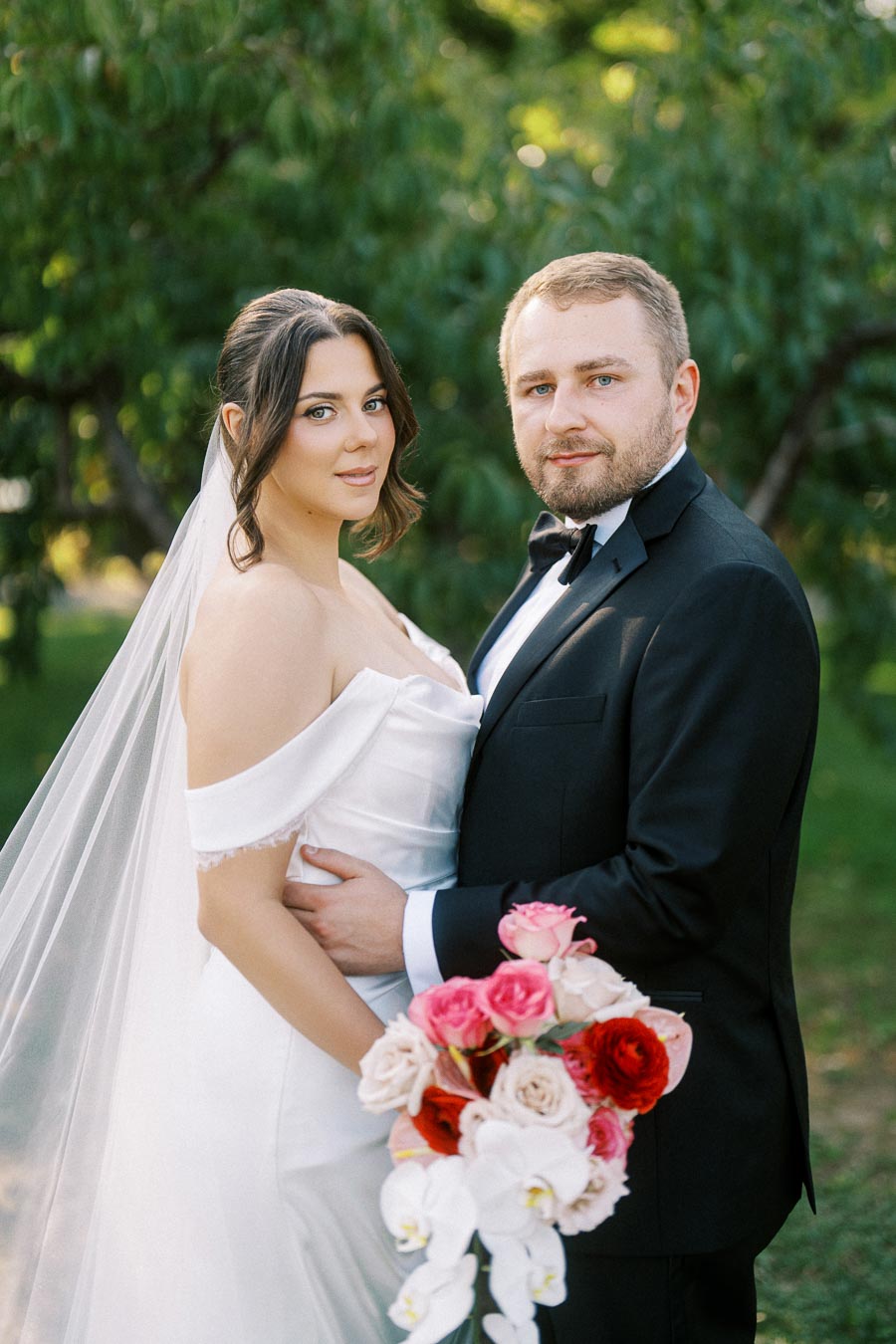 A bride and groom pose romantically in a lush green garden. The bride is wearing an elegant off-shoulder white wedding gown and veil, holding a vibrant bouquet of pink and red flowers. The groom is dressed in a classic black tuxedo with a bow tie.