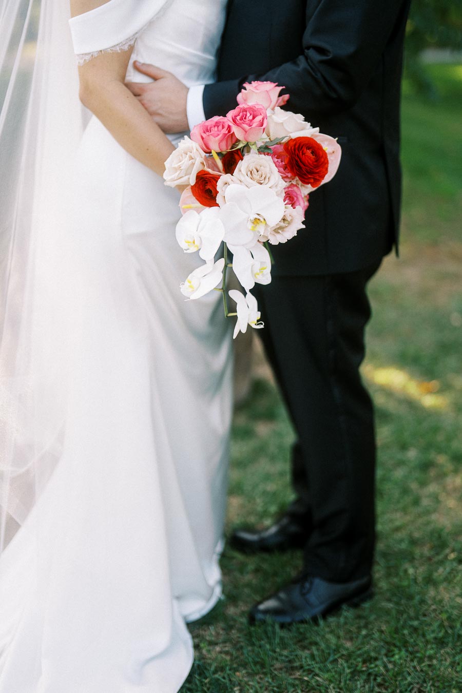 Bride and groom embracing outdoors with bouquet of red, pink, and white flowers, featuring roses and orchids.