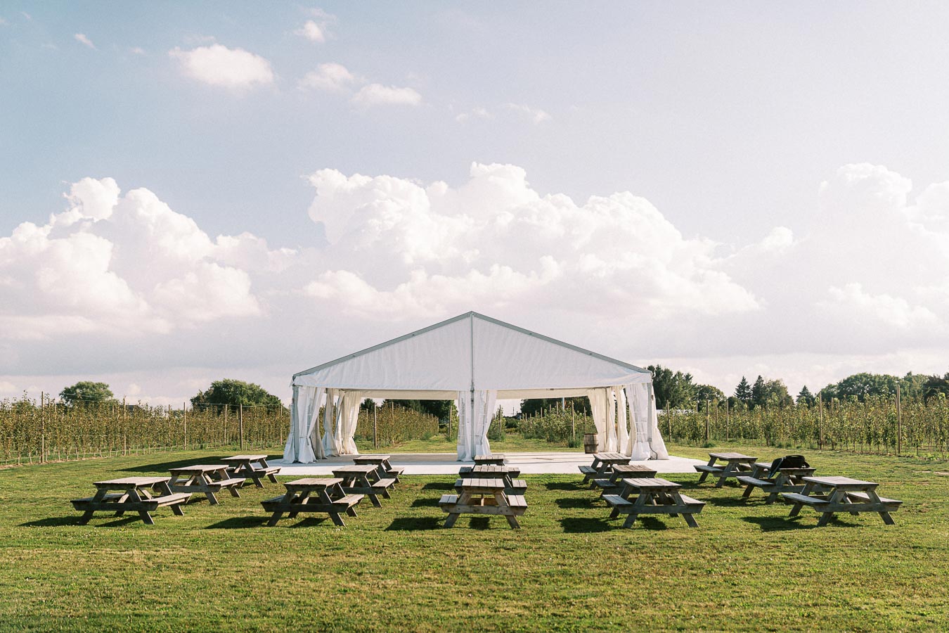 Outdoor event setup with a white tent surrounded by picnic tables on a grassy field under a partly cloudy sky.