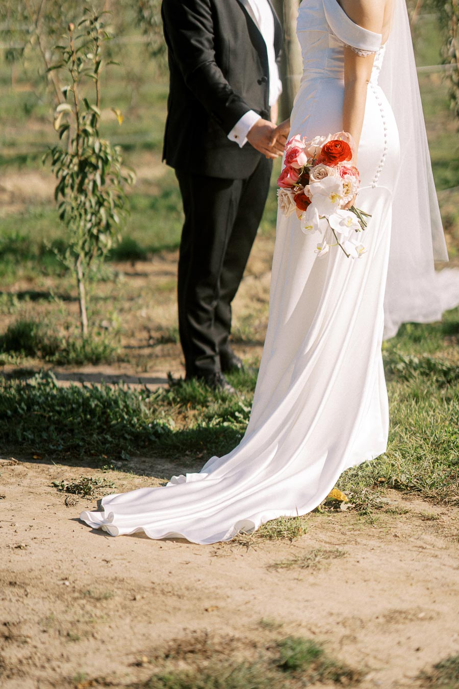 Bride in elegant white wedding dress holding a bouquet of red and white flowers, standing next to groom in black suit, surrounded by lush greenery in outdoor setting.