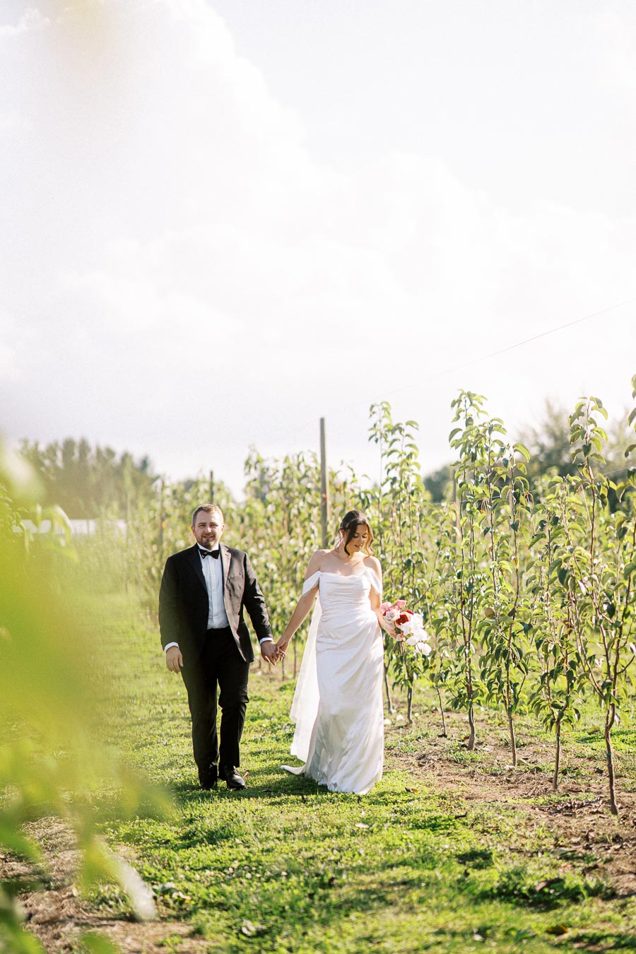 Wedding couple walking hand in hand through a lush orchard, with the bride in a beautiful white gown holding a bouquet of flowers and the groom in a black tuxedo, under a bright, sunny sky.