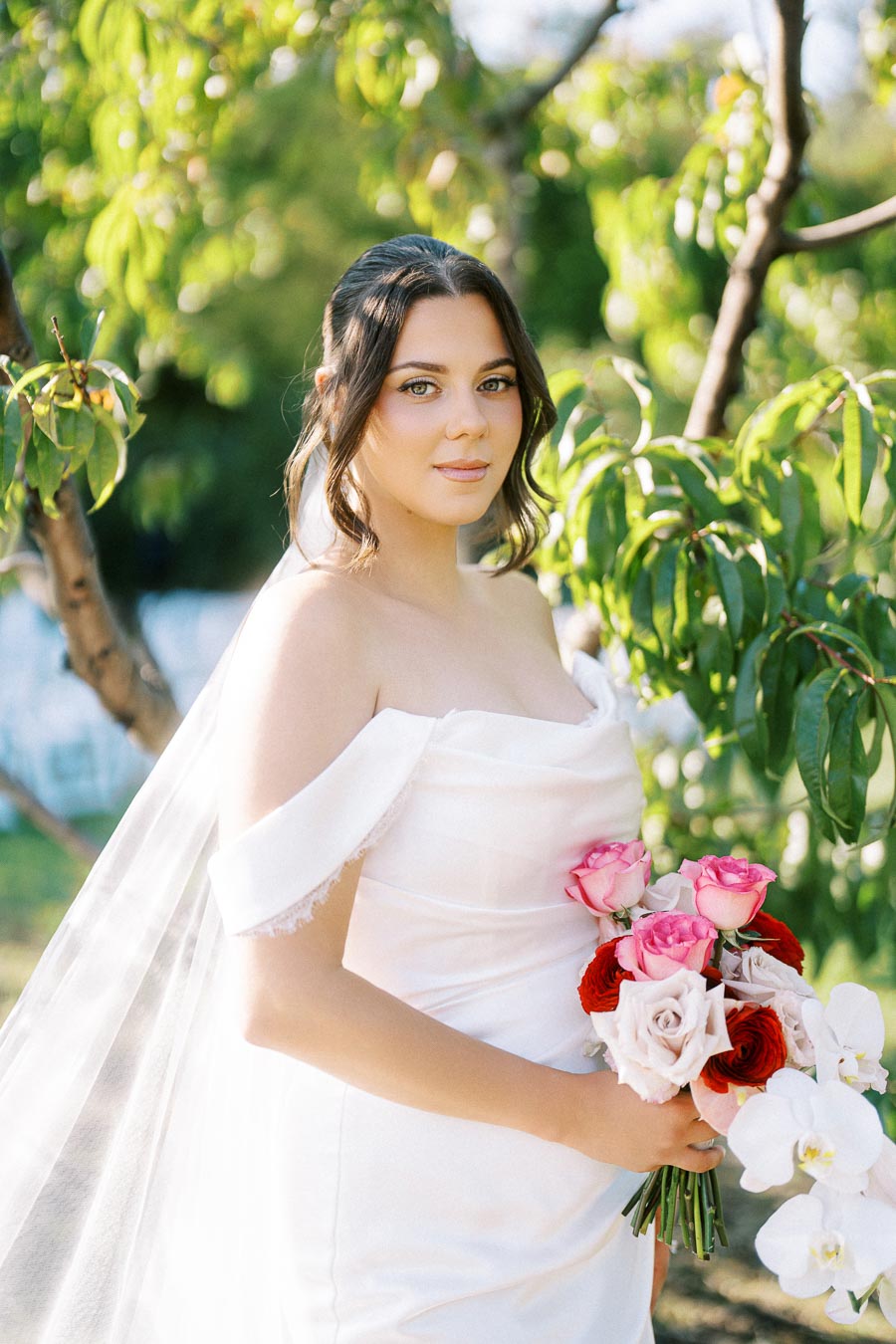 Bridal portrait of a woman in an elegant white gown holding a vibrant bouquet of pink roses and orchids, set against a lush outdoor backdrop with green foliage.