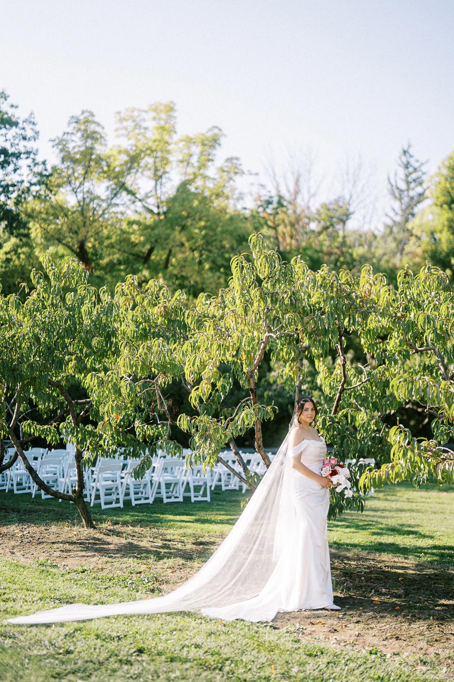 A bride in a flowing white dress with a long veil stands outdoors amidst lush green trees, holding a vibrant bouquet, with rows of white folding chairs in the background, suggesting a garden wedding setting.