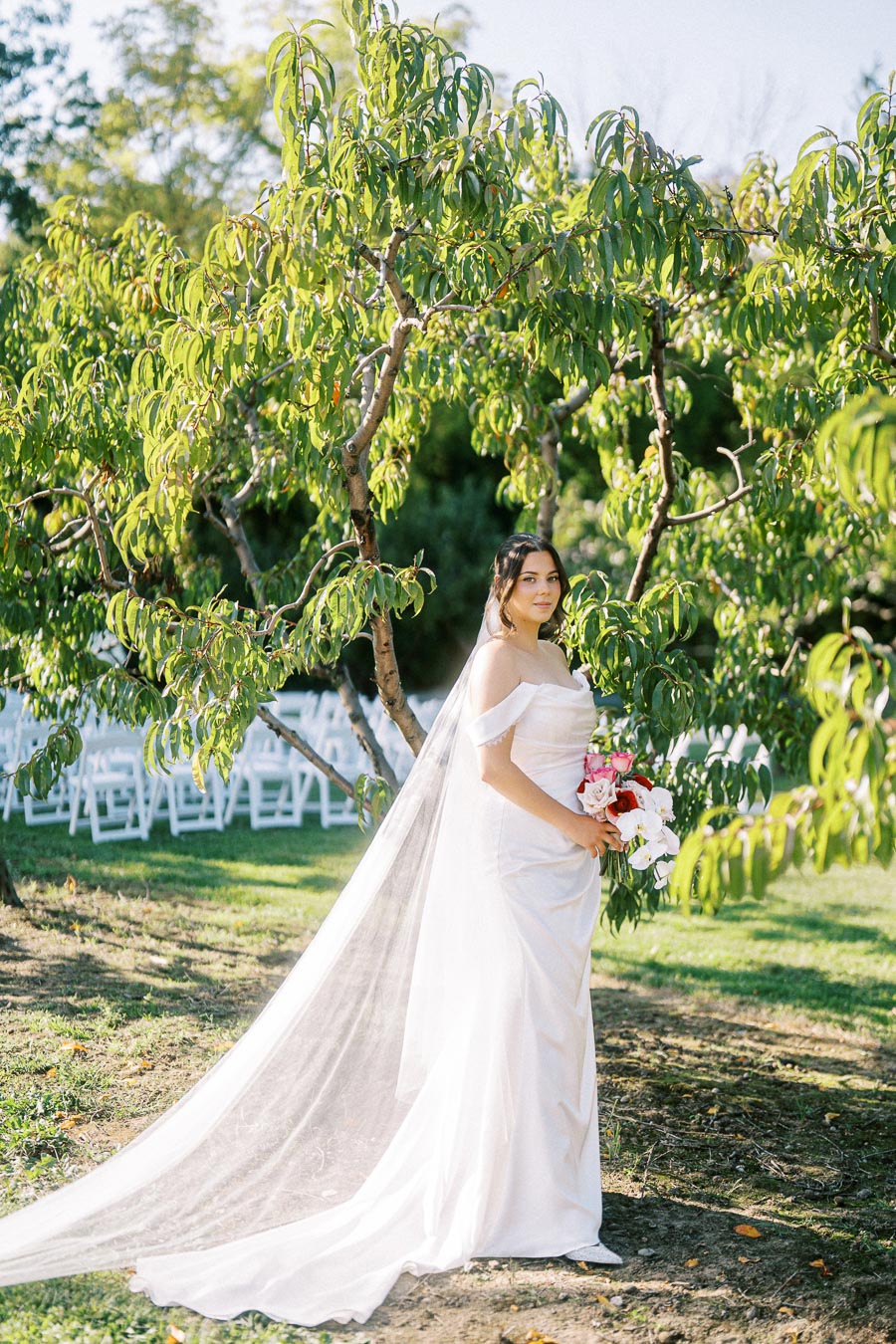 Elegant bride in an off-shoulder white wedding gown with a long veil, holding a vibrant bouquet, stands gracefully in a lush garden setting with green trees and white chairs in the background.
