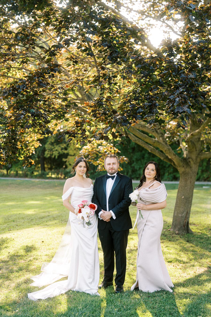 Bride and groom with bridesmaid standing in a sunlit garden, surrounded by lush greenery and a large tree, all dressed in elegant formal attire for a wedding ceremony. The bride holds a bouquet of red and white flowers, while the bridesmaid holds a single white rose.