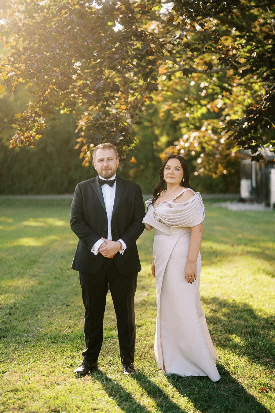 Elegant couple dressed in formal attire standing on a sunlit grassy lawn, with trees and golden leaves in the background.
