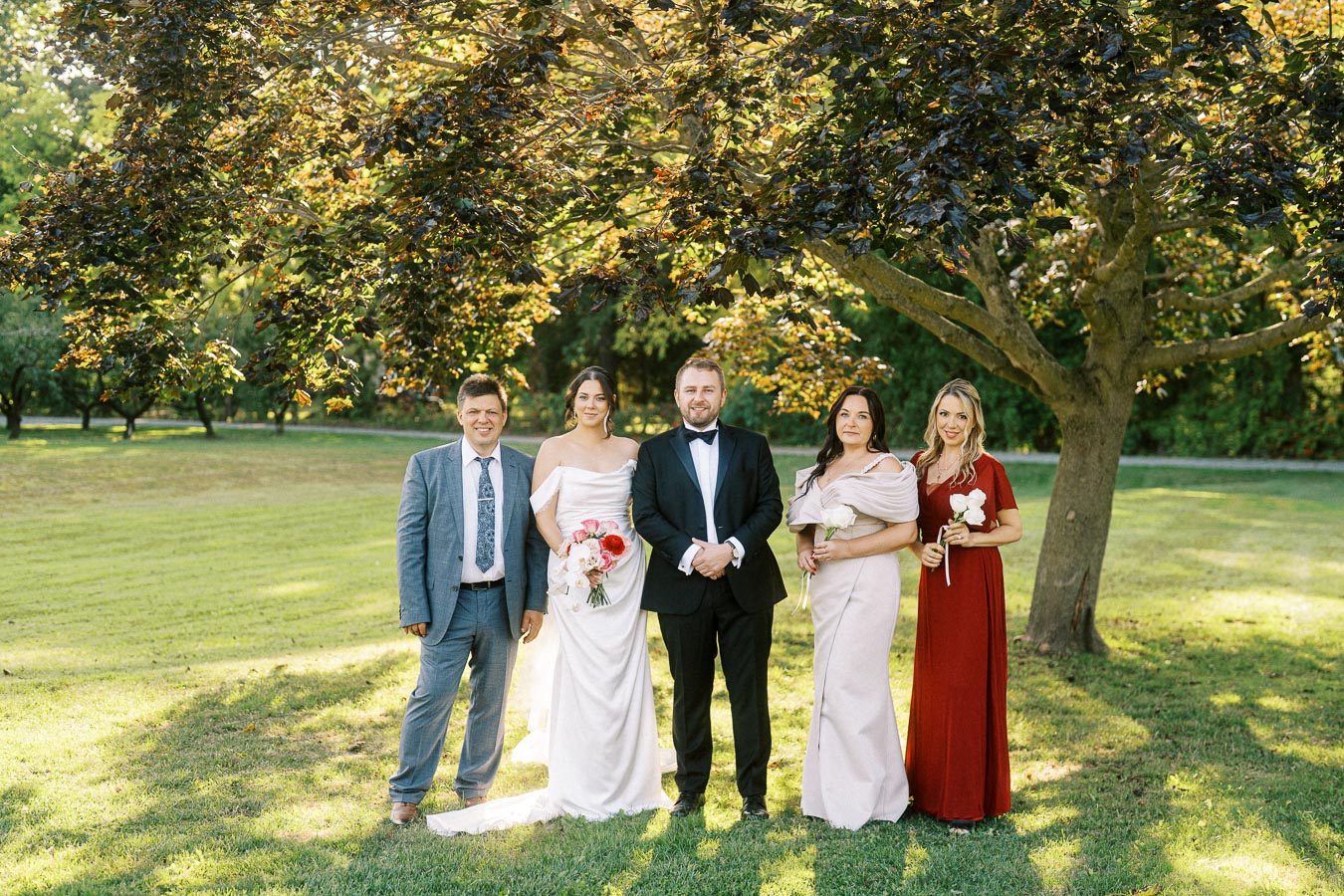 A wedding party stands together under a large tree, featuring a bride in a white gown holding a bouquet of pink and white flowers, a groom in a black tuxedo, and three well-dressed individuals in suits and elegant dresses on a sunny day.