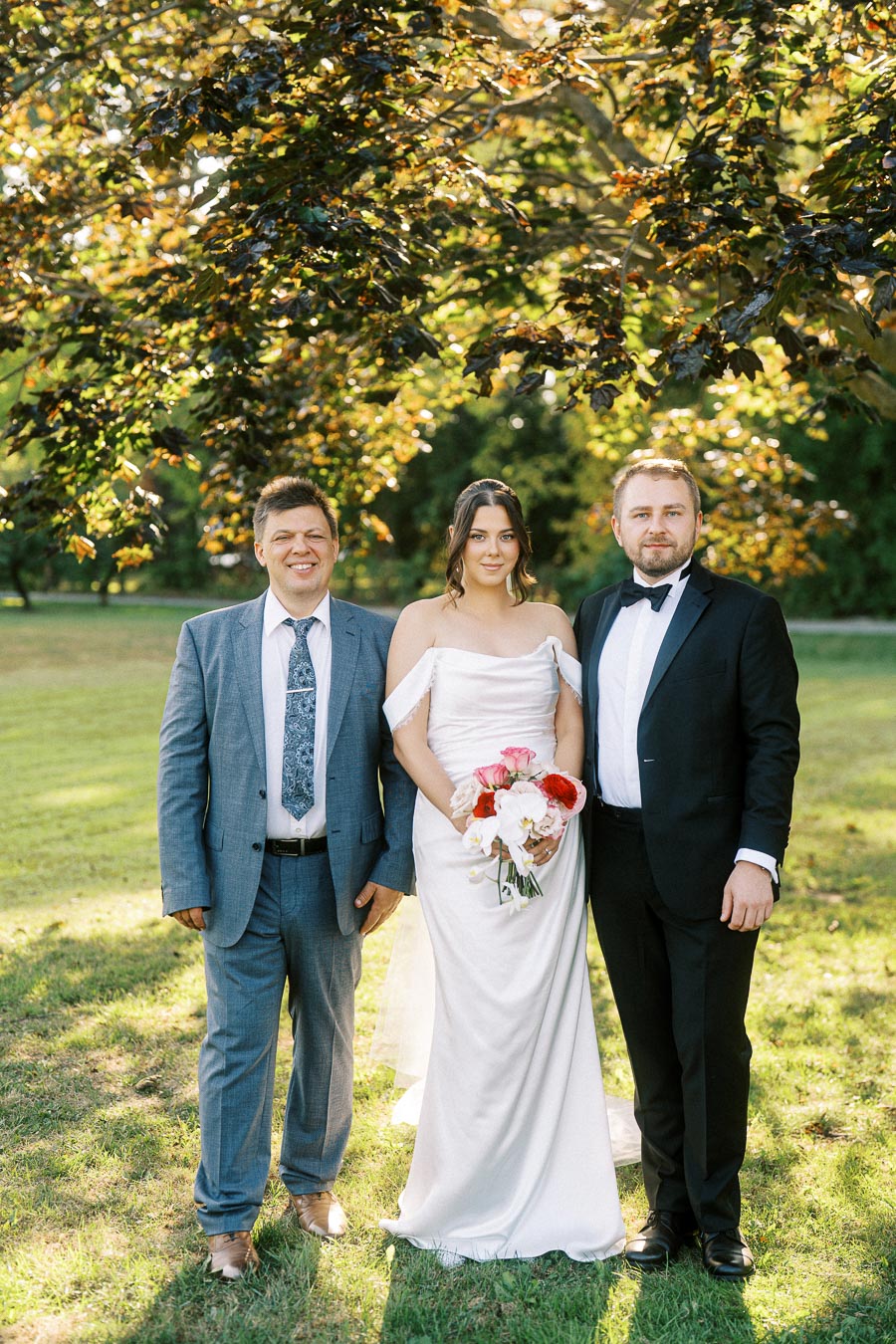 A bride in a white gown holding a bouquet stands between two men in suits under a tree with sunlight filtering through the leaves, outdoor portrait on a wedding day.