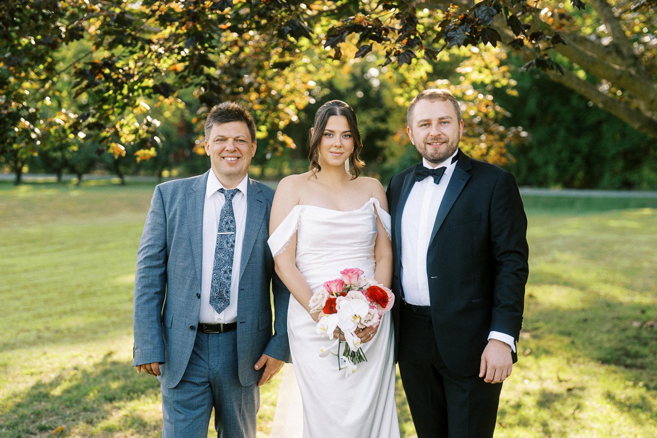A bride in a white gown holding a bouquet, standing between two men in suits, outdoors under a tree with green foliage.