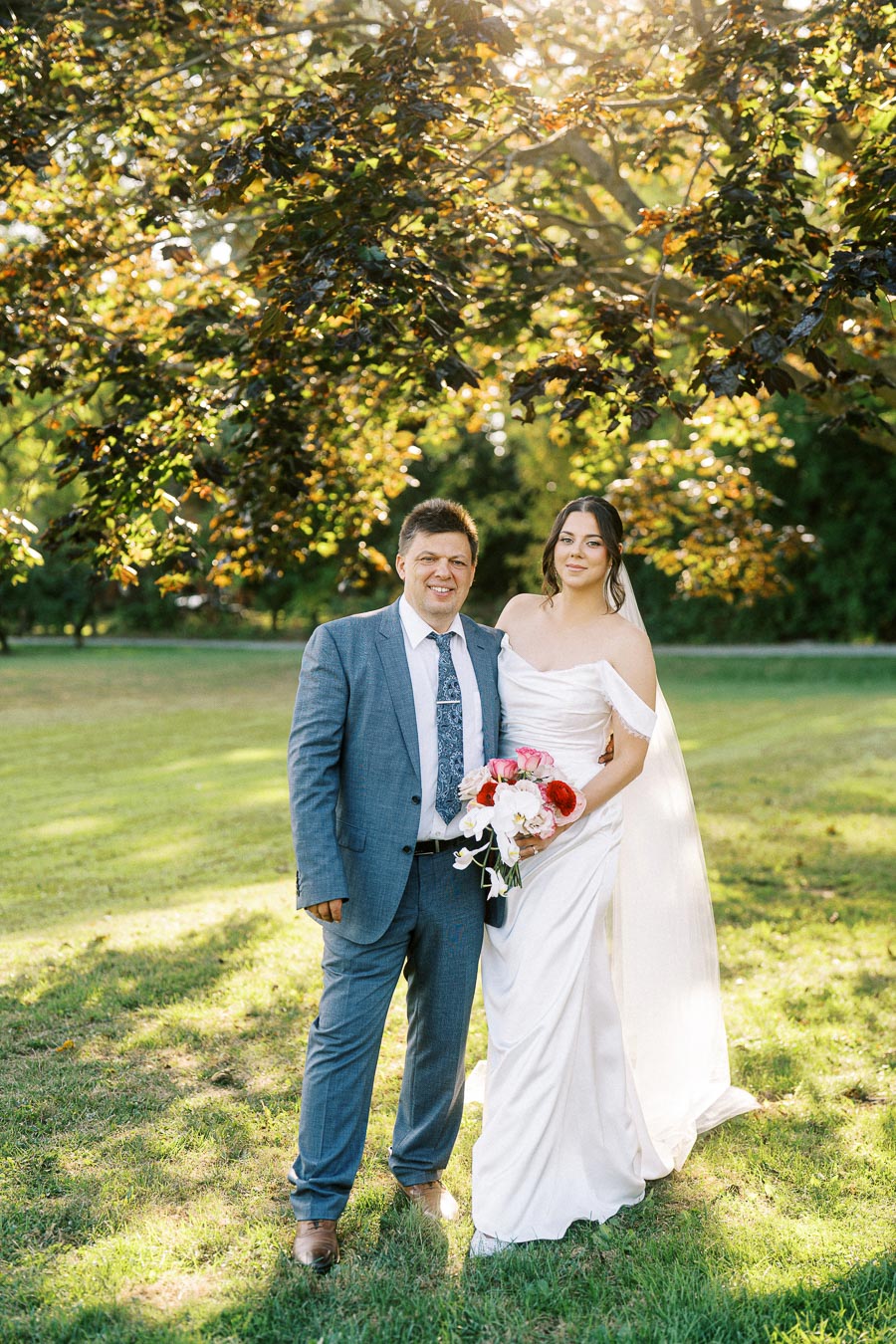 Smiling bride and groom posing outdoors in lush green garden beneath a tree during a sunny wedding day
