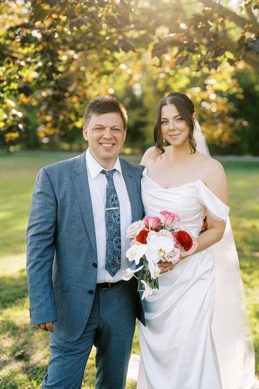 A bride in a white dress holding a bouquet of colorful flowers poses with a man in a blue suit in a sunny outdoor setting, surrounded by green foliage.
