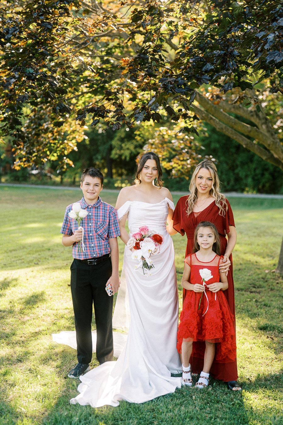 A bride in a white dress holds a bouquet of flowers, standing with three children under a tree in a sunlit garden. The children are dressed in red and plaid, holding flowers, smiling. Perfect day for an outdoor wedding celebration.