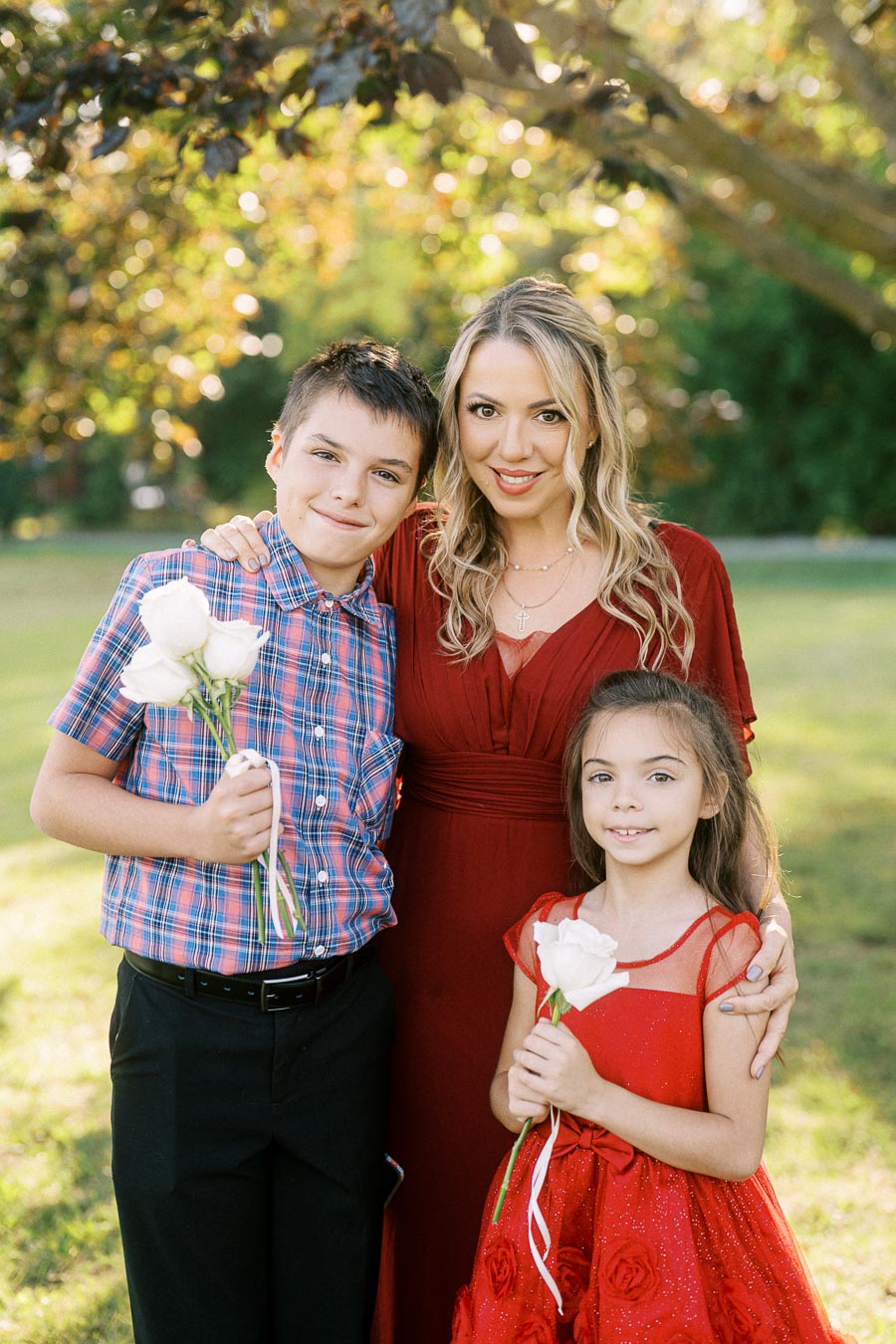 A woman in a red dress stands outside with two children, a boy holding white roses and a girl in a red dress, against a backdrop of green trees and sunlight.