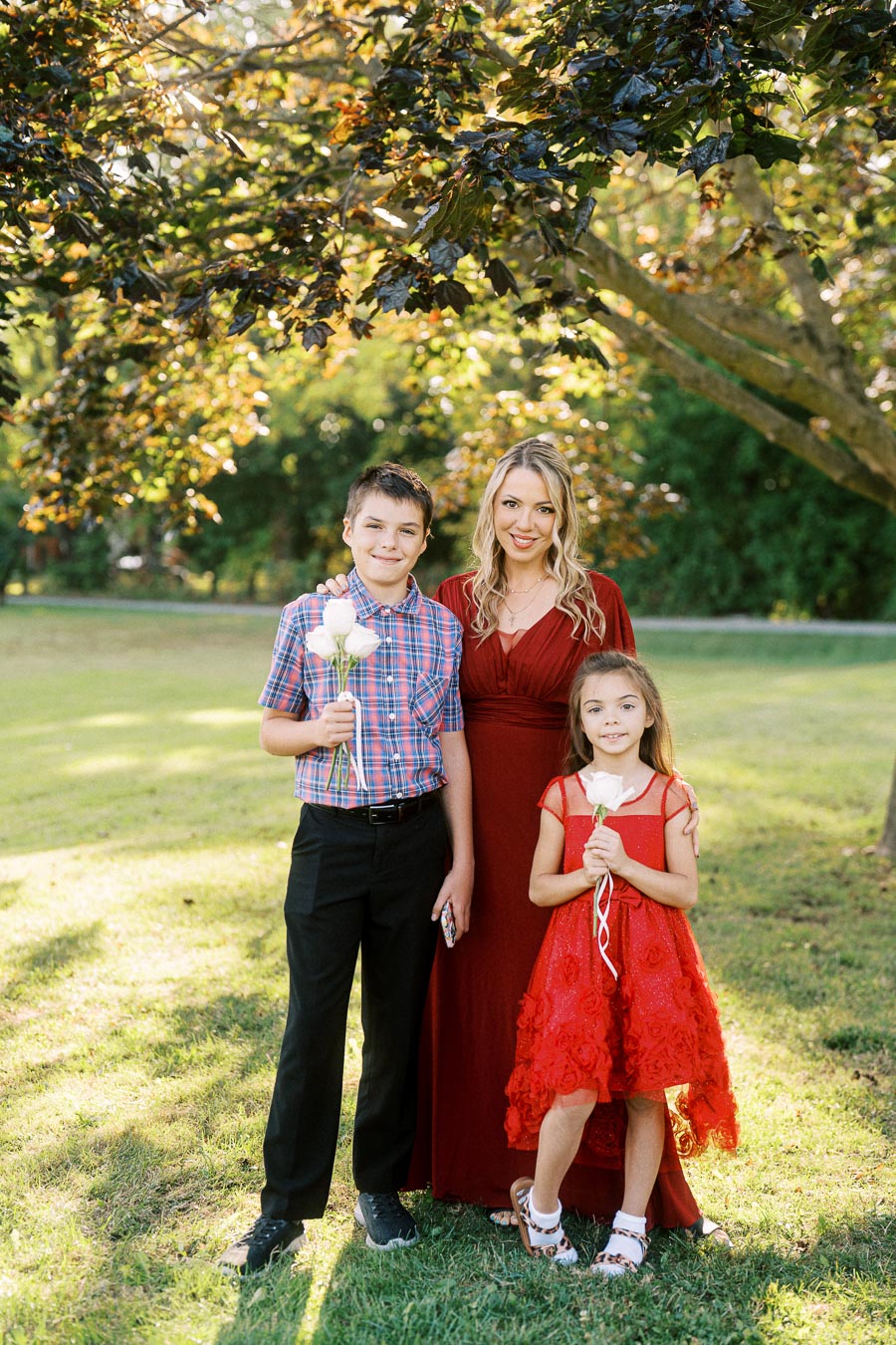 A woman in a red dress poses with two children holding flowers under a tree on a sunny day, set in a green park.