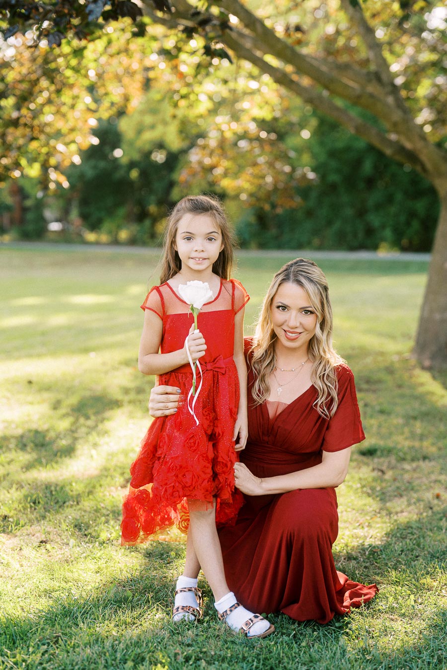 A woman and a young girl, both in elegant red dresses, pose together in a sunlit park. The girl is holding a white rose, and the lush green trees create a serene background.
