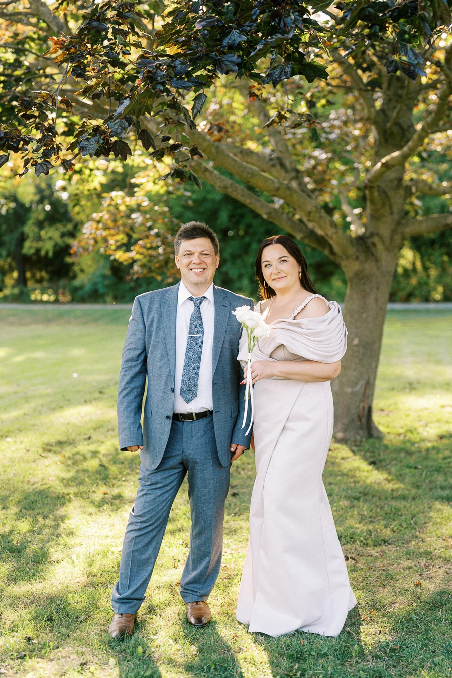 A couple standing in a sunlit park, the man in a blue suit and tie, and the woman in an elegant beige dress with a bouquet of white flowers, posing under a large tree with green foliage.