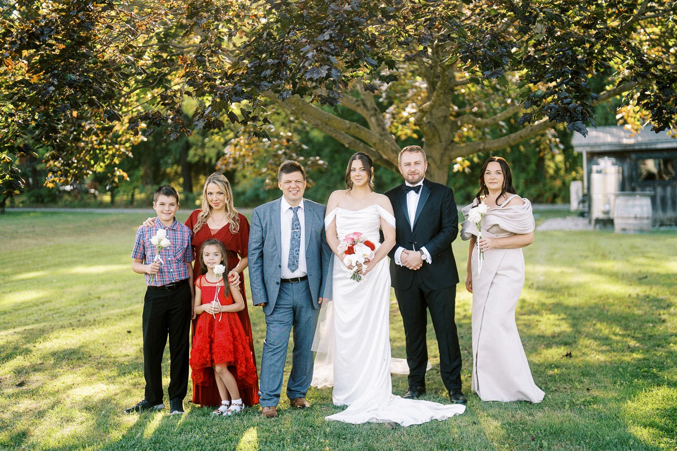 A wedding party poses outdoors under a large tree on a sunny day. The bride in a white dress and groom in a tuxedo stand in the center, surrounded by family and friends dressed in formal attire. The green grass and natural setting provide a picturesque backdrop.