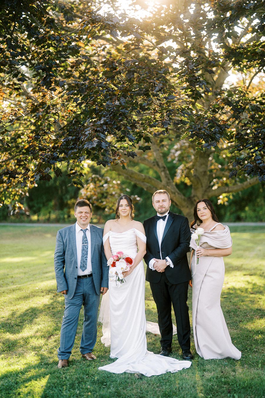 A wedding party of four people standing outdoors under a tree, with the bride in a white gown and holding a bouquet of flowers, the groom in a black tuxedo, and two guests in formal attire, all smiling in a sunlit garden setting.
