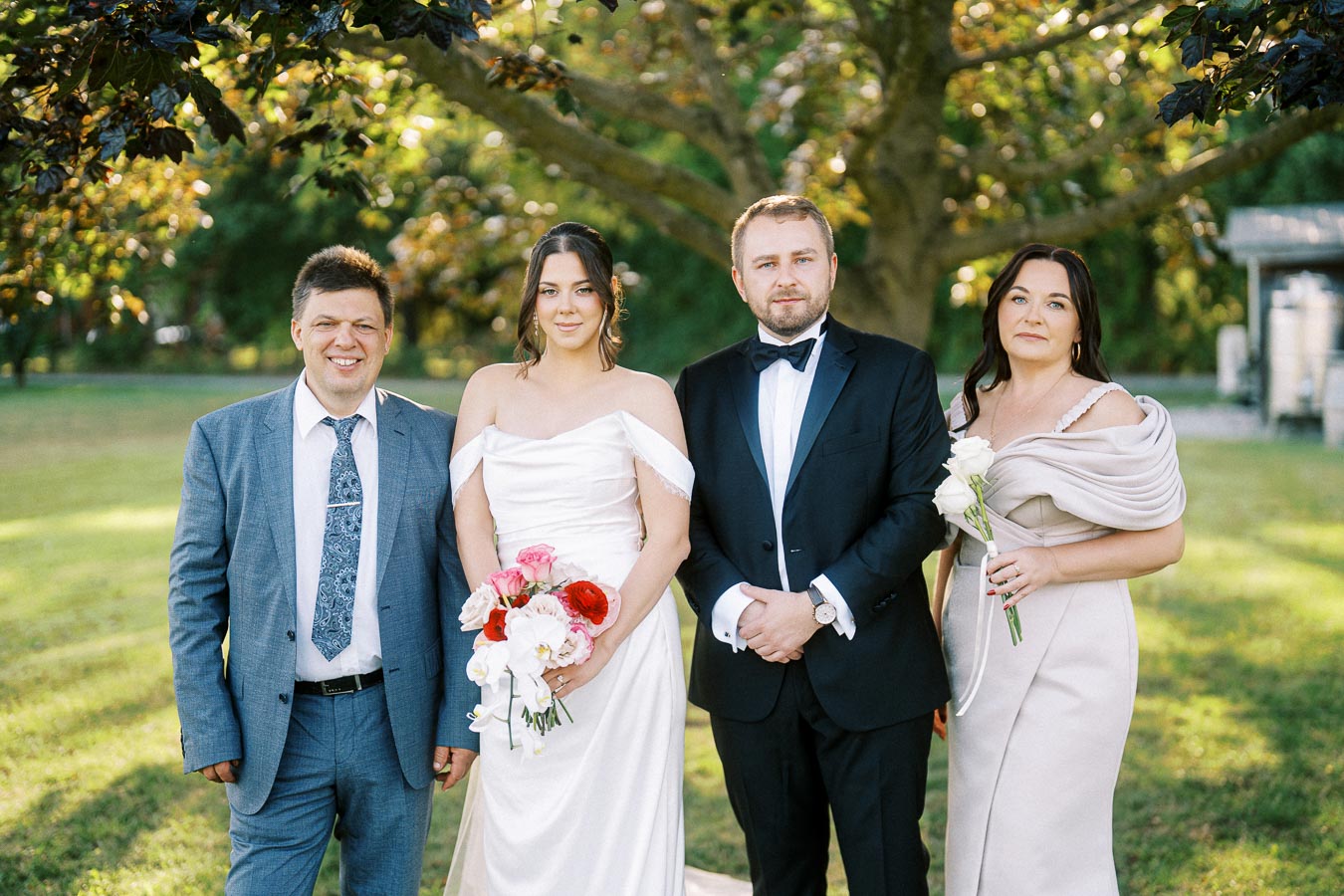 Wedding group photo in a garden setting with four people, including a bride in a white dress holding a colorful bouquet, a groom in a black tuxedo, and two elegantly dressed guests.