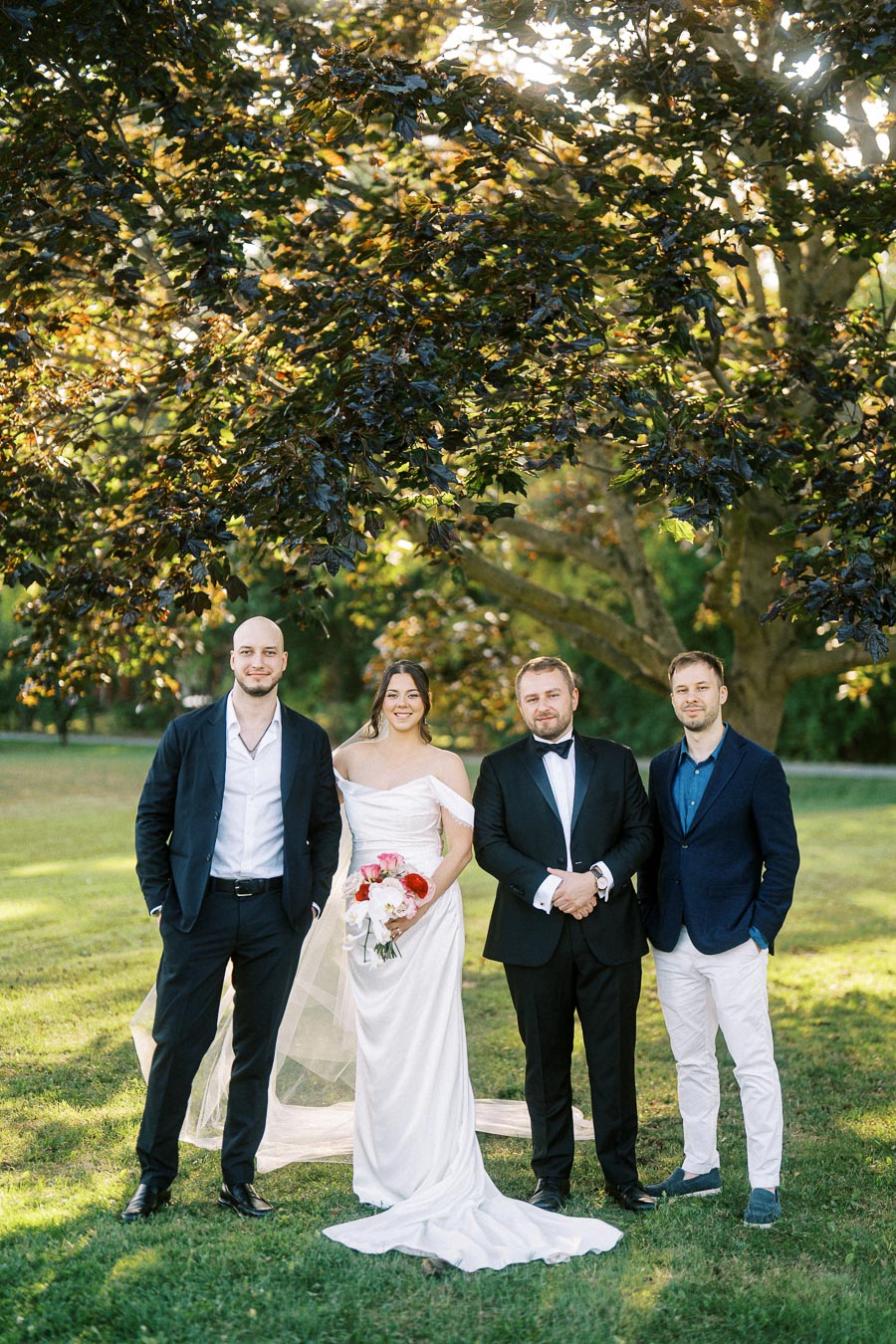 Wedding group photo with a bride in an elegant white gown holding a bouquet, standing alongside three groomsmen in formal suits, under a large tree in a lush green park setting.