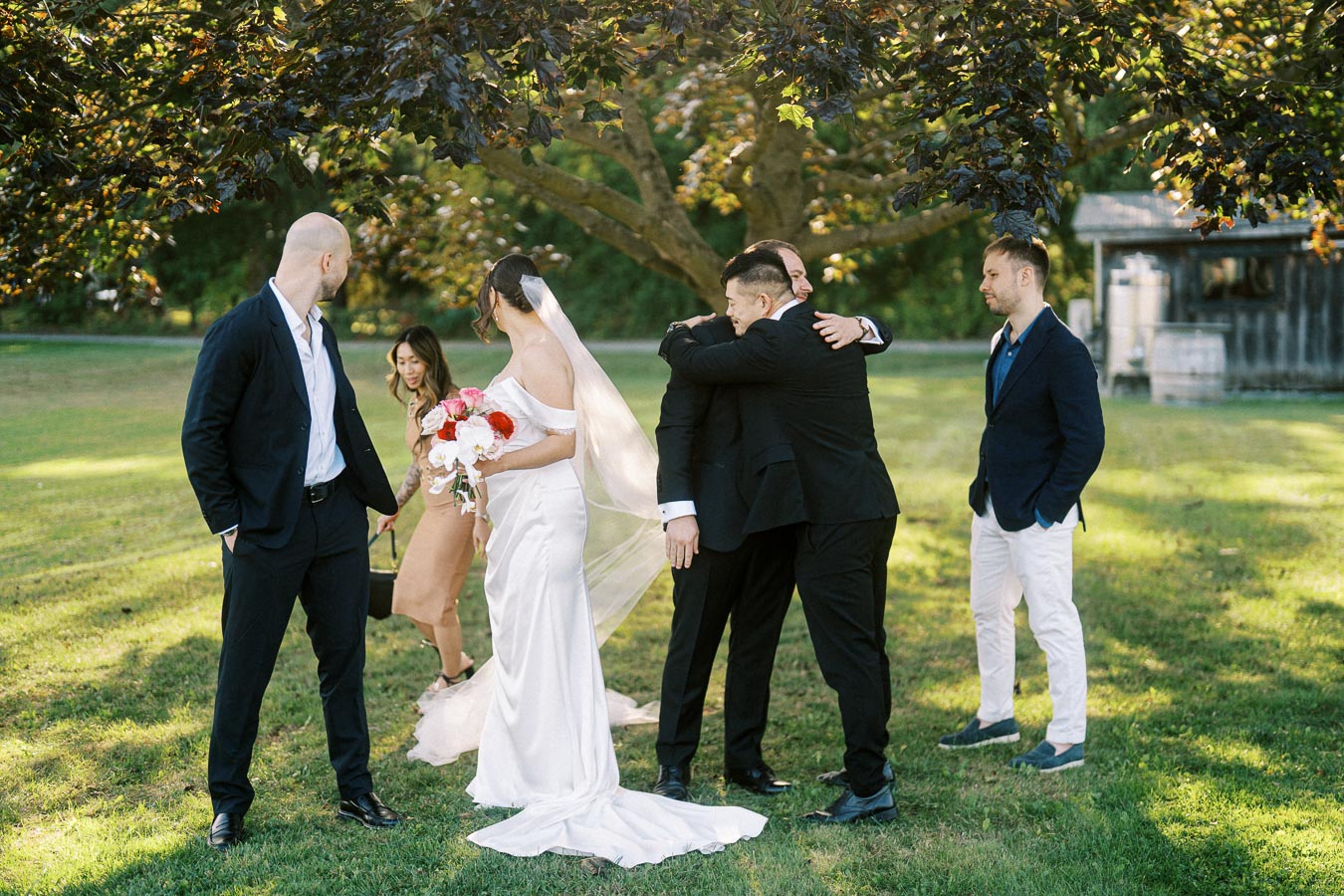 A bride in a white wedding gown holds a bouquet of pink and red flowers, standing on a grassy lawn under a large tree. She interacts with guests dressed in formal attire, capturing a joyful moment at an outdoor wedding gathering.