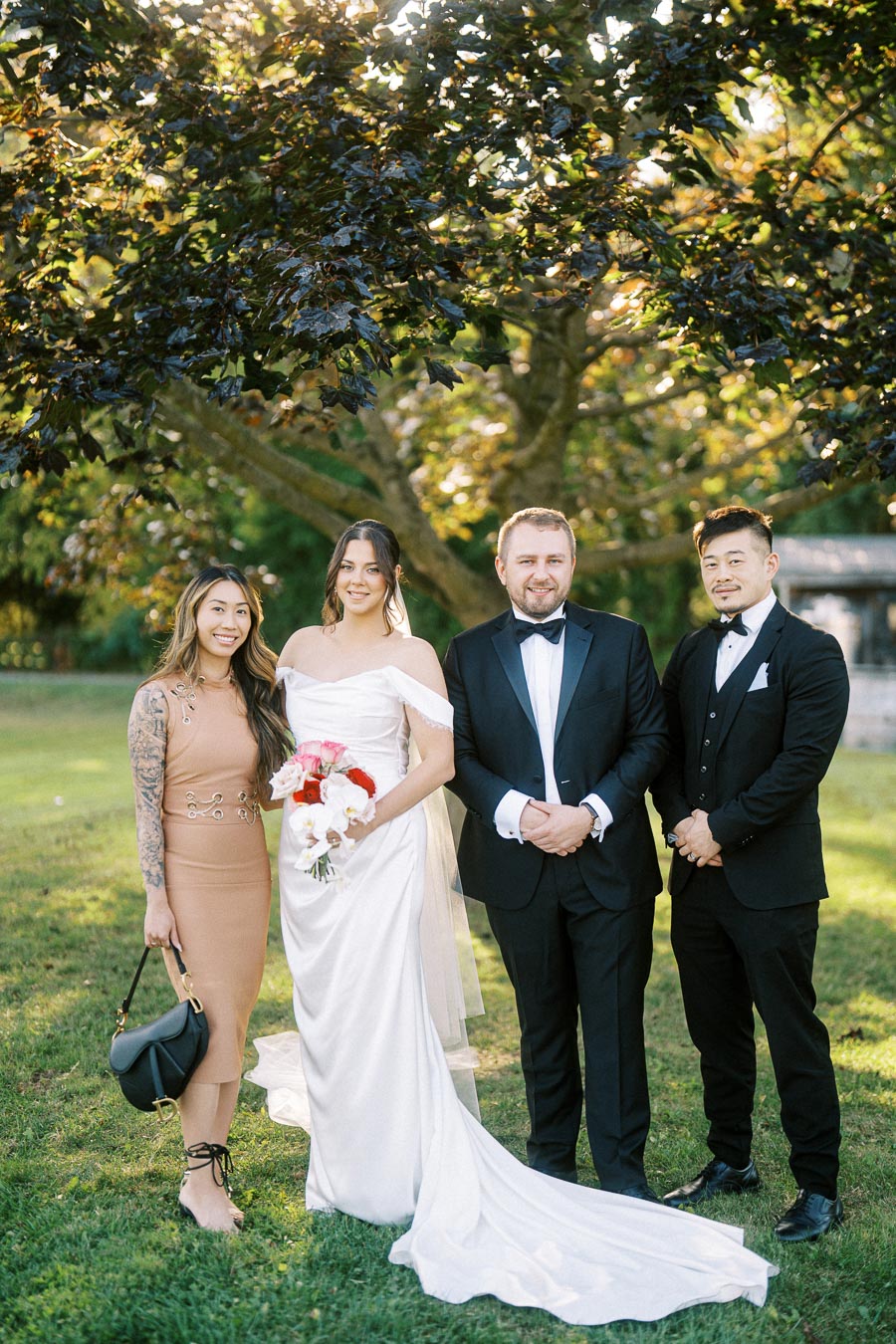 A bride in a white wedding dress stands with three guests, two men in suits and a woman in a tan dress, under a tree in a sunlit garden.