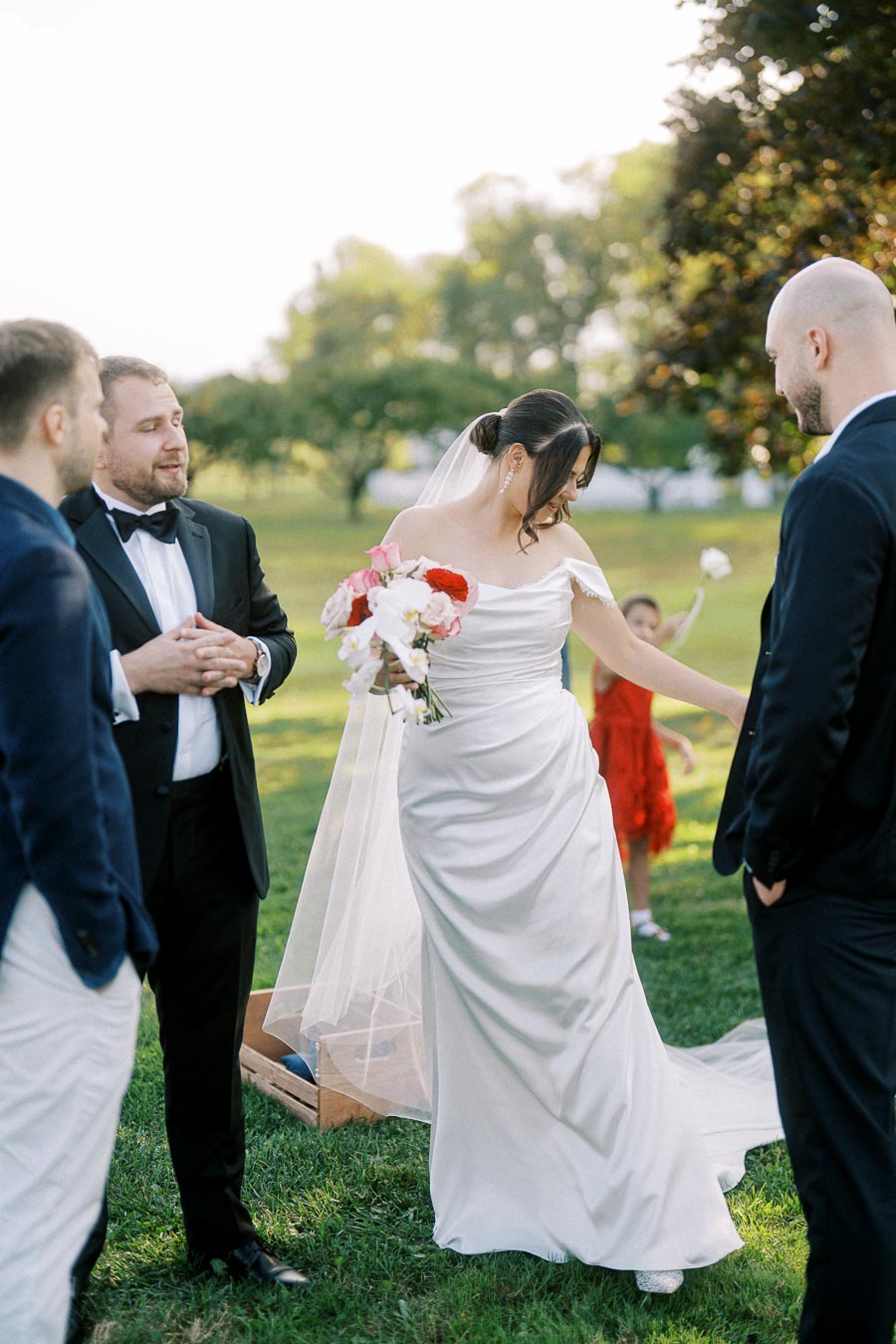 A bride in a white wedding dress holding a bouquet of red and white flowers, smiling and interacting with guests in formal attire on a grassy lawn during an outdoor wedding ceremony.