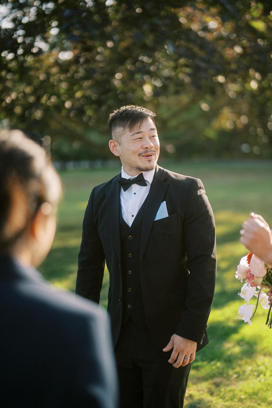A person wearing a black suit with a bow tie smiling outdoors at a wedding ceremony, holding a bouquet. Sunlight filters through surrounding greenery, creating a warm and joyful atmosphere.