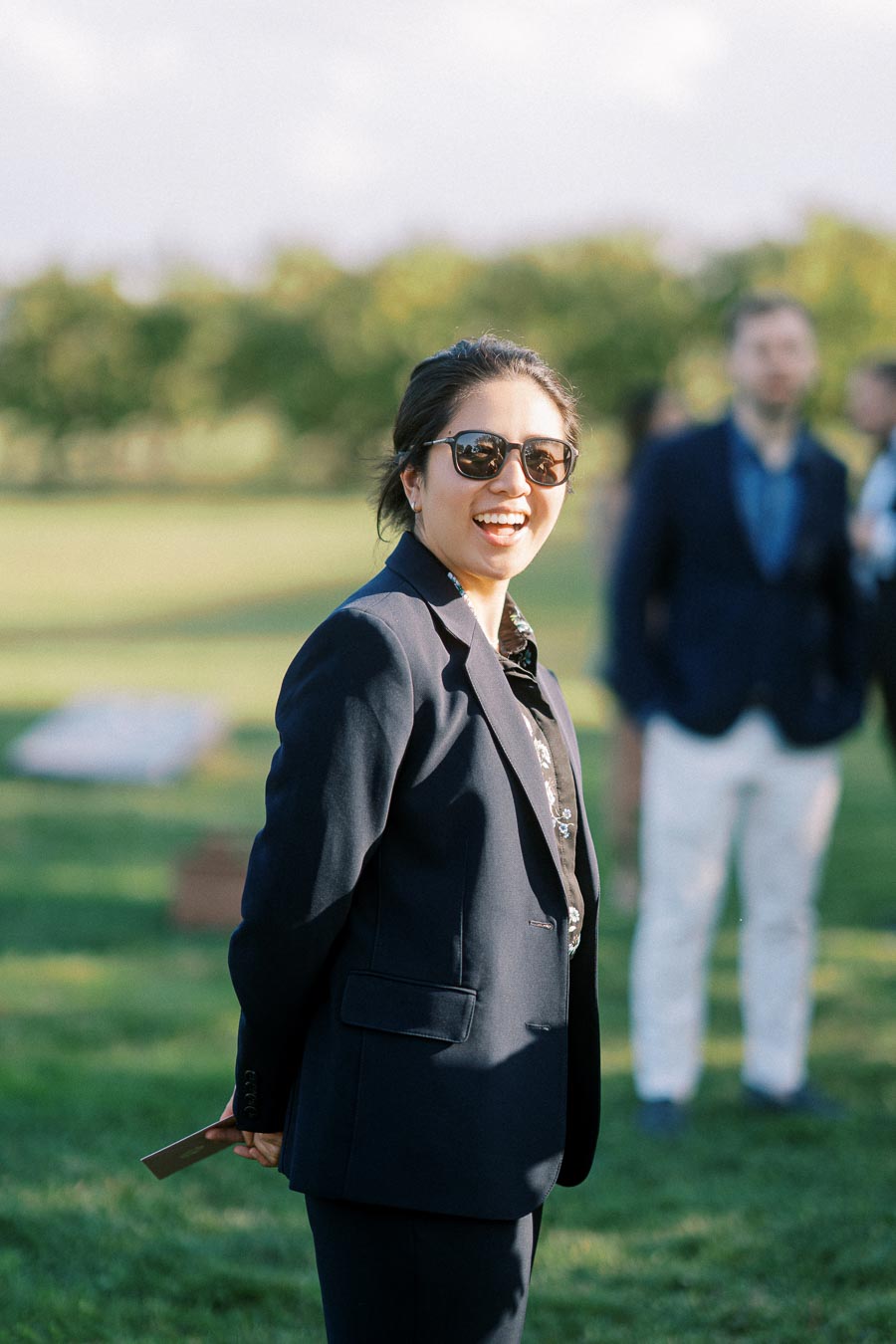 A person wearing sunglasses and a navy blazer smiles in a sunny outdoor setting with blurred people and greenery in the background.