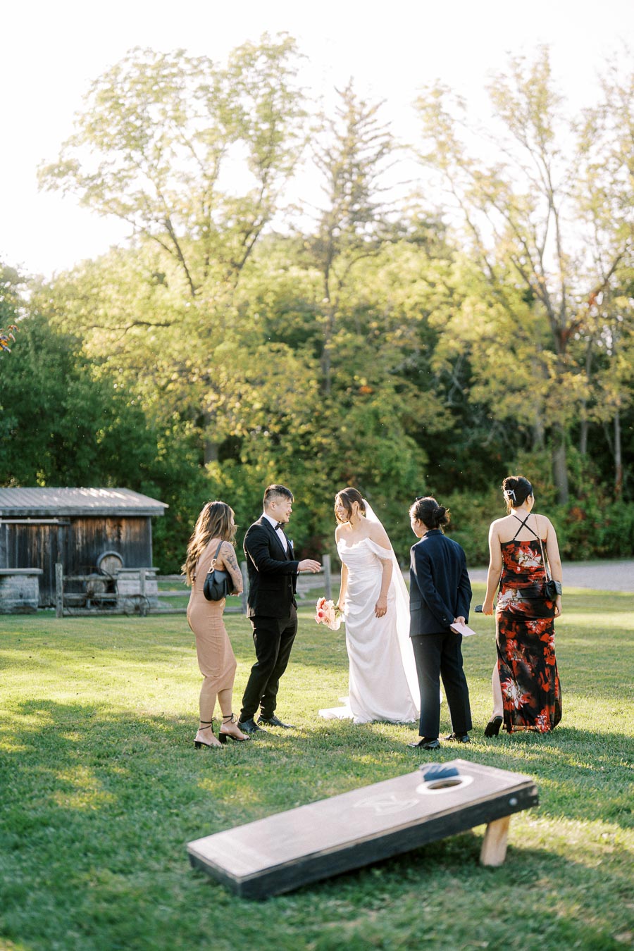 A small group of people, including a bride in a white gown holding a bouquet, engage in conversation on a green lawn with trees and a rustic wooden barn in the background. A cornhole game board is visible in the foreground, capturing a casual outdoor wedding celebration scene.