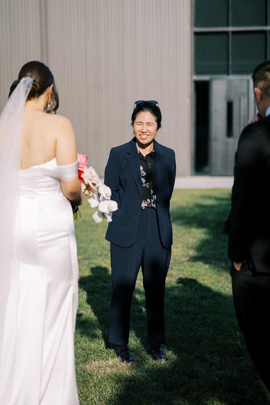 A bride in a white wedding dress and veil holds a bouquet, standing outdoors on a sunny day, interacting with guests wearing formal attire.
