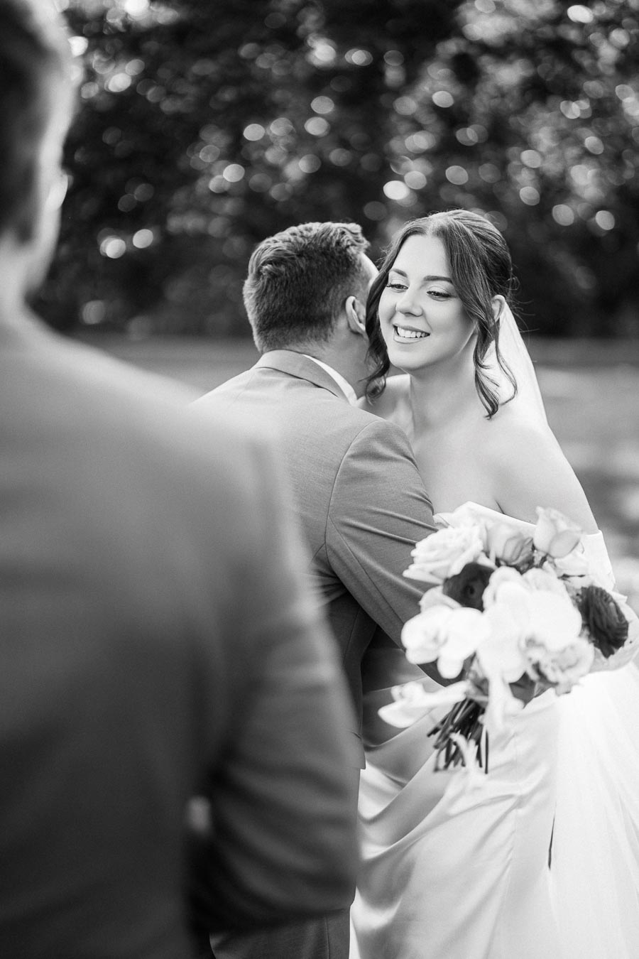 Black and white image of a joyful bride holding a bouquet, smiling while hugging a groom in a suit during an outdoor wedding ceremony.