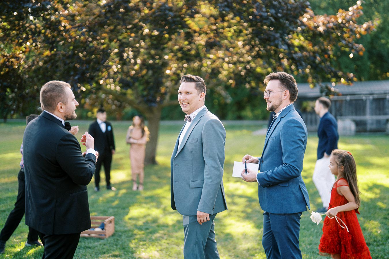A group of people in formal attire enjoying an outdoor event with a lush garden backdrop, featuring a joyful conversation among guests and a young girl in a red dress holding a white flower.