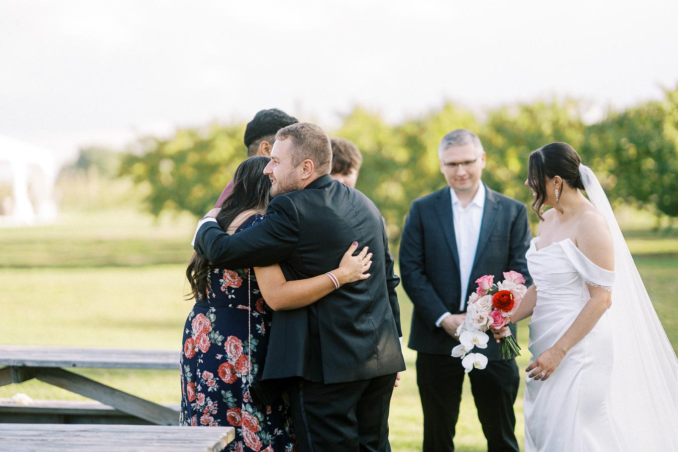 A bride in a white dress holds a vibrant bouquet at an outdoor wedding, as a man in a suit hugs a guest wearing a floral dress. A few attendees stand nearby on a sunny day with greenery in the background.