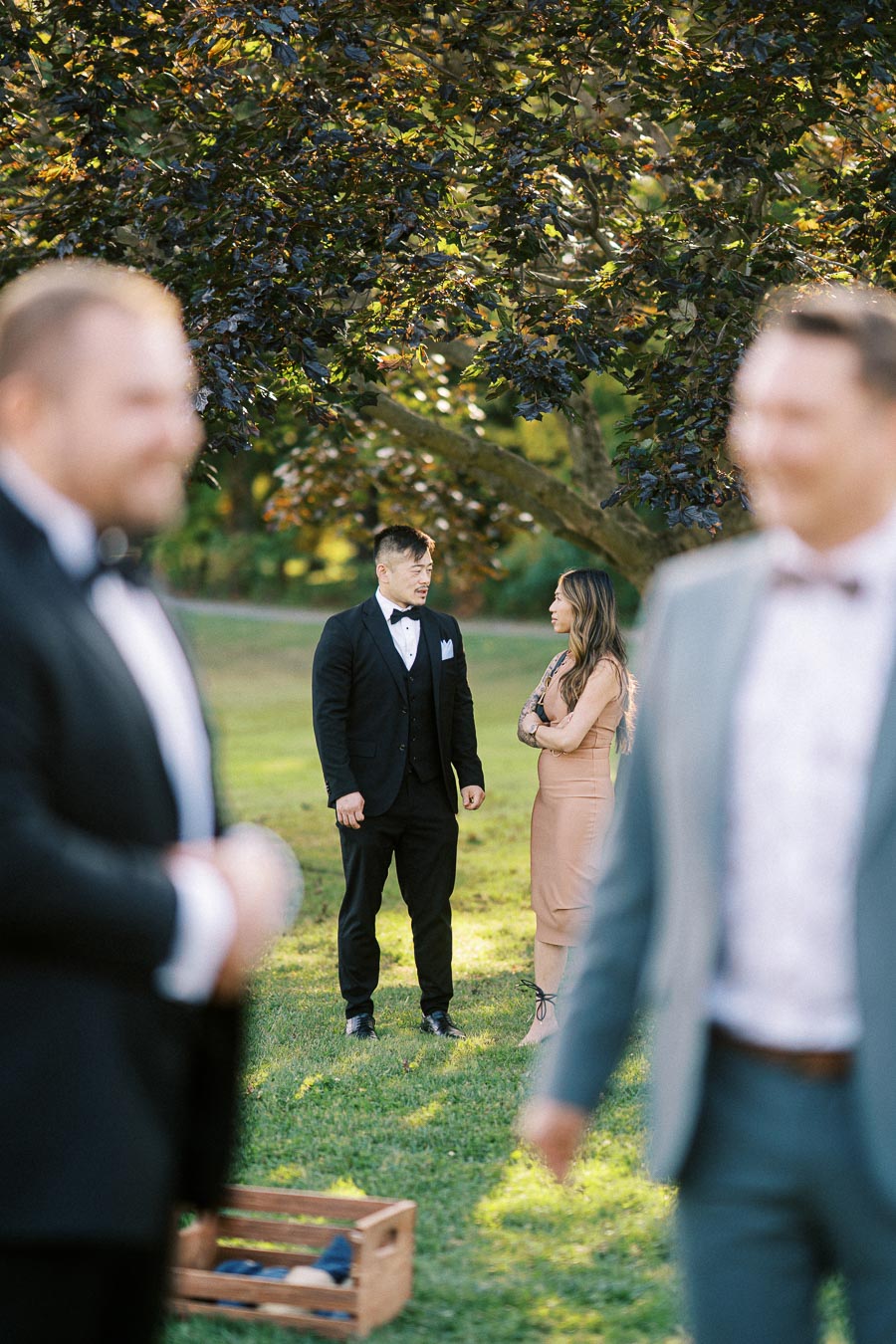 Wedding guests socializing outdoors in elegant attire, with a focus on a well-dressed man and woman talking under a tree.