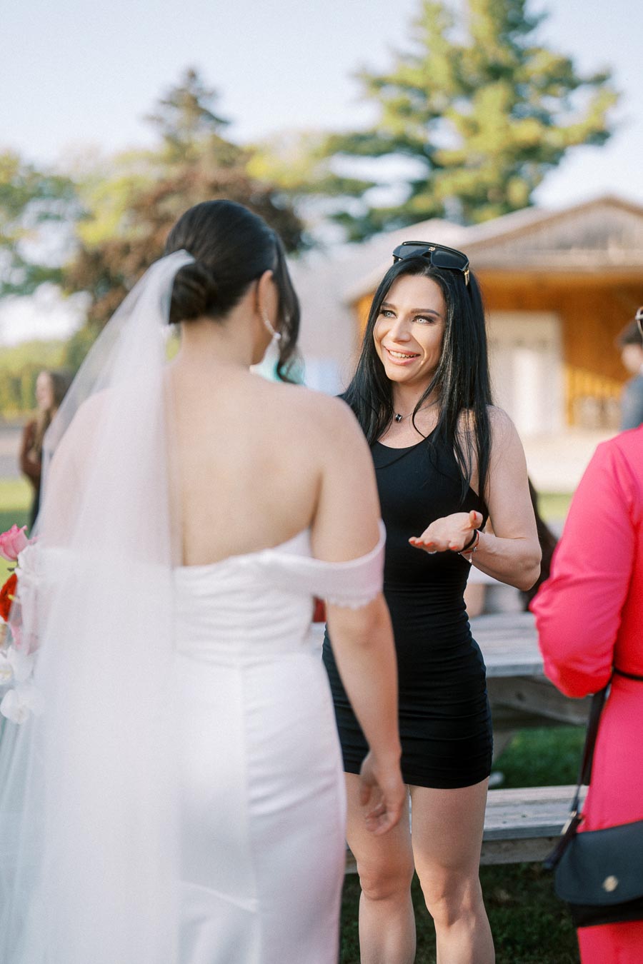Two women happily conversing outdoors at a wedding; one is a bride in a white gown, and the other is wearing a black dress.