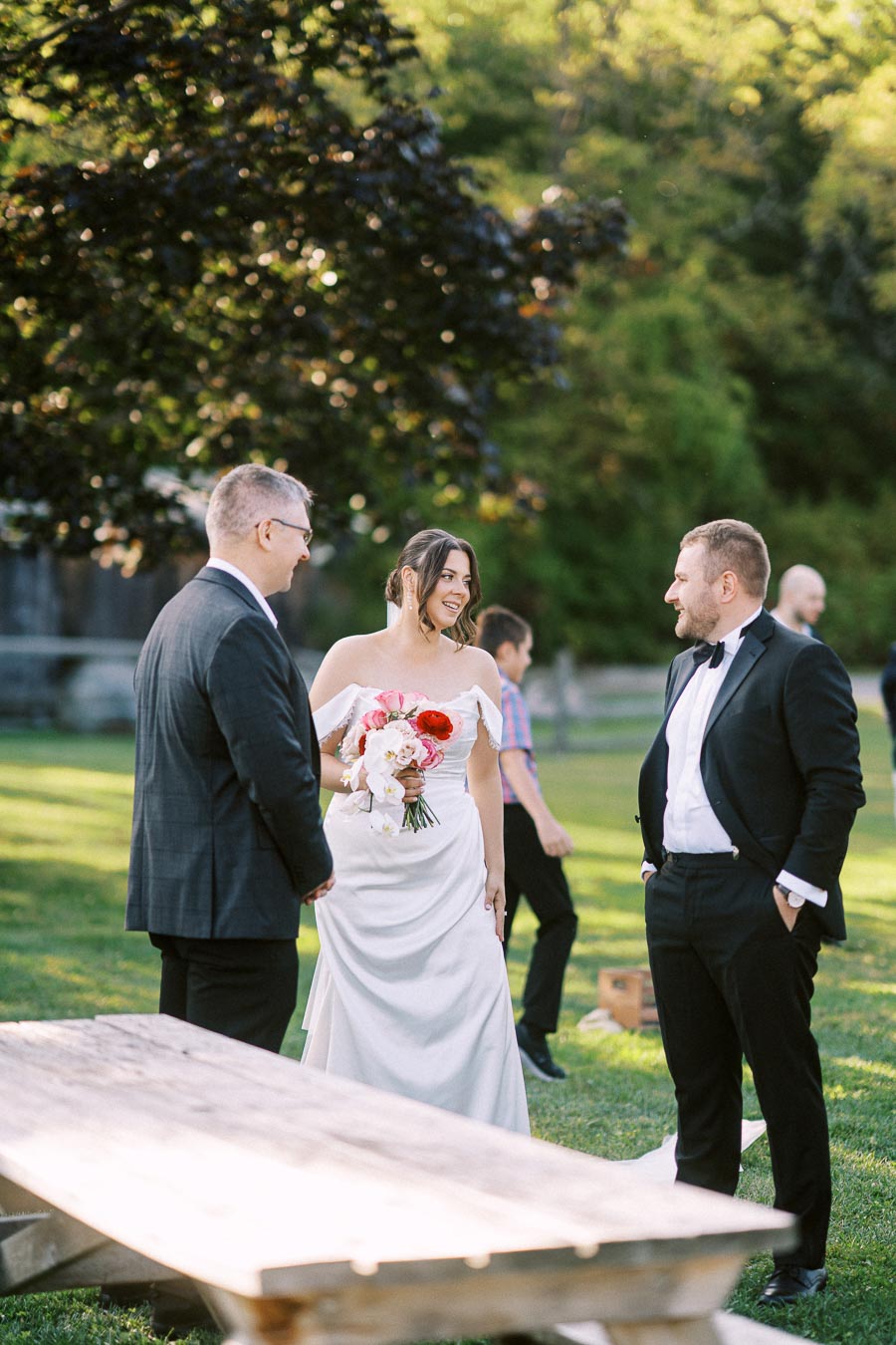 A bride in a white wedding dress holds a bouquet while smiling and conversing with two men in formal attire at an outdoor wedding reception, surrounded by green trees and a child walking nearby.