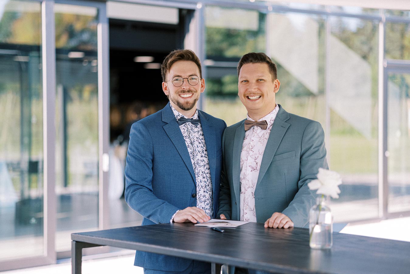Two people in formal suits with floral shirts and bow ties standing and smiling at a table outdoors.