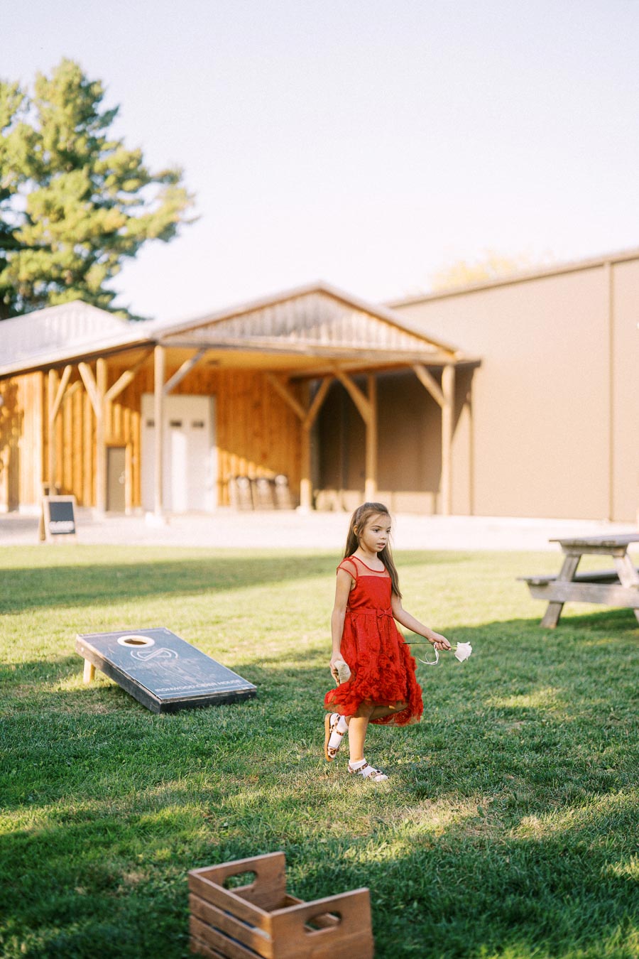 A young girl in a red dress walks across a grassy lawn, holding a flower, with a rustic wooden building and picnic bench in the background, and a bean bag toss game set up on the grass.