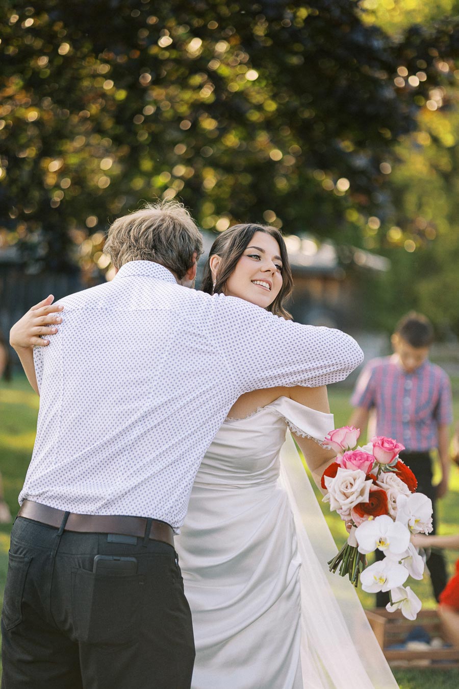 A bride smiling and holding a bouquet of colorful flowers while sharing a heartfelt hug at an outdoor wedding ceremony, with a blurred background of greenery and guests.