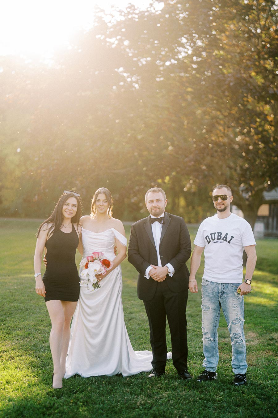 A bride and groom are standing with two friends in a sunlit garden, capturing a joyful moment on their wedding day; the bride holds a bouquet of flowers, while the groom is in a tuxedo.