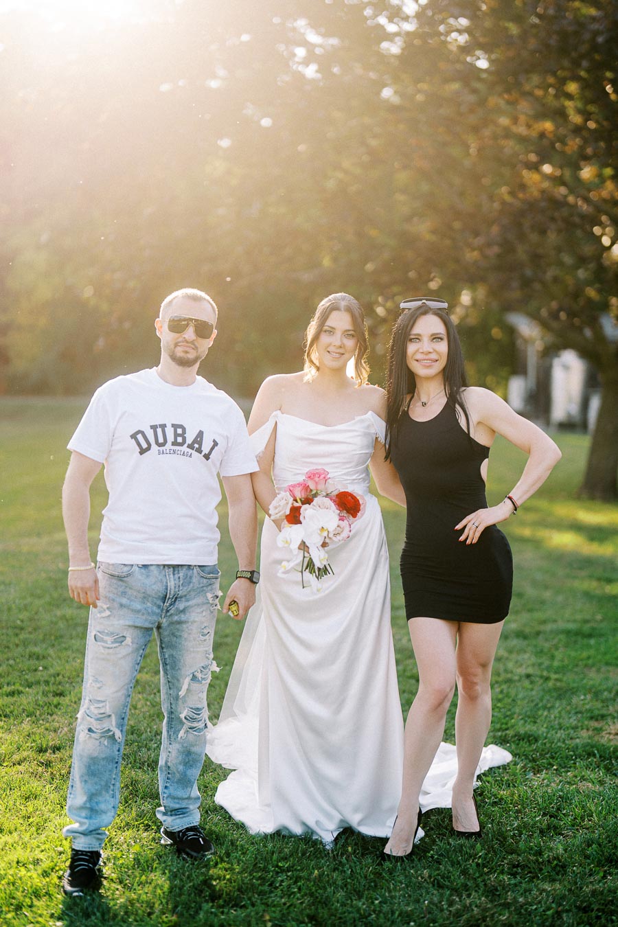 A bride in a white dress holding a colorful bouquet stands between two friends outdoors in a sunlit garden, with one friend in casual attire and the other in a sleek black dress.