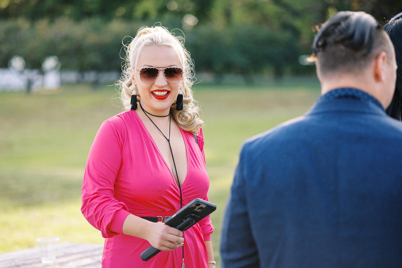 A woman in a bright pink dress and sunglasses smiling while holding a phone, standing outdoors on a sunny day.