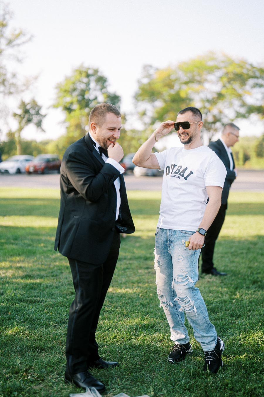 Two men are laughing outdoors on a grassy area. One is dressed in a formal suit, while the other is wearing casual attire with sunglasses, a white t-shirt, and ripped jeans. The background features trees and parked cars, creating a relaxed and joyful atmosphere.