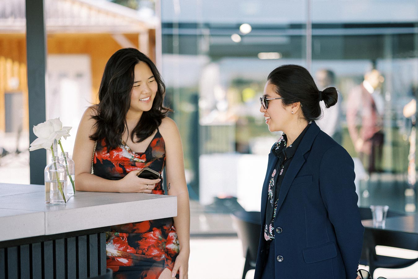 Two women having a friendly conversation outdoors, one in a colorful floral dress holding a smartphone, the other in a navy blazer with sunglasses, standing by a modern cafe counter with white roses in a vase.