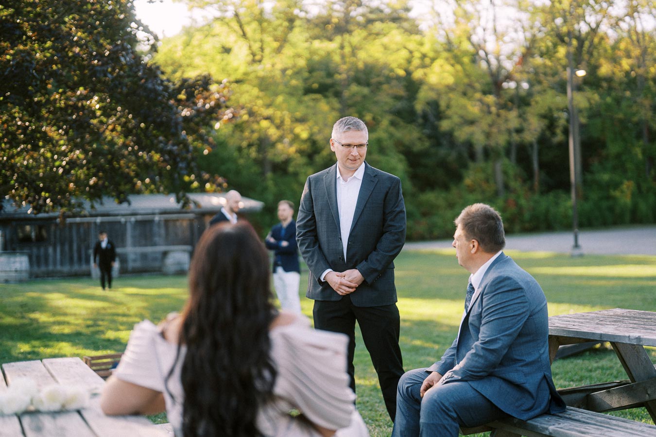 A group of people in formal attire having a conversation outdoors on a sunny day, with picnic tables and trees in the background, suggesting a relaxed, professional gathering.
