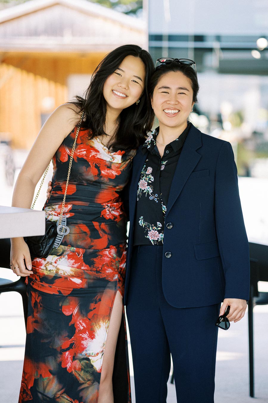 Two people smiling and posing together, one in a floral red dress and the other in a navy suit with floral patterns, standing outside a modern building with a wooden structure in the background.
