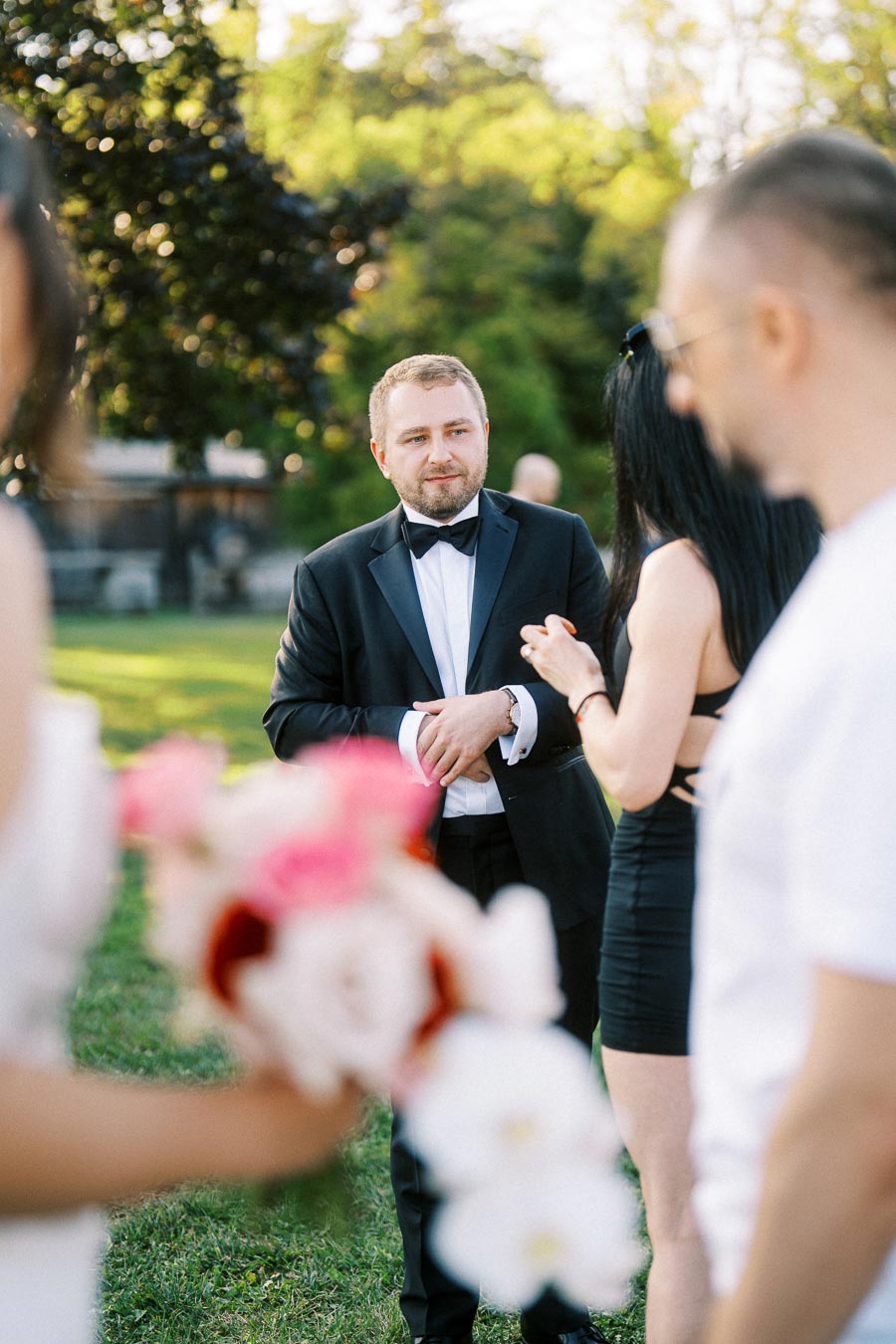 A man in a black tuxedo stands outside on a grassy lawn during a formal event, surrounded by people holding bouquets of flowers.